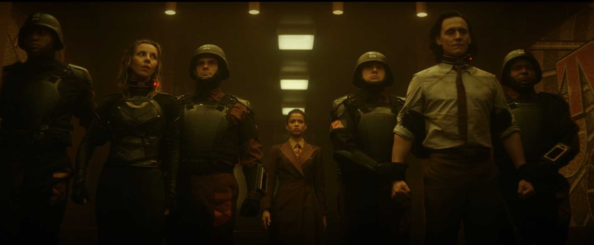 Loki and Sylvie are captured by TVA soldiers, in all black with helmets. Sylvie is on the left, wet hair, Loki outfit. Loki is on the right, TVA outfit, flexing his arms against the two soldiers that restrain him. In the background we see Ravonna looking somber."